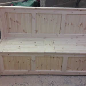 May include: A light-coloured wooden bench with a high backrest and a storage compartment underneath. The piece features a panelled design with vertical slats on both the backrest and the front base, showcasing the natural grain and knots of the wood.