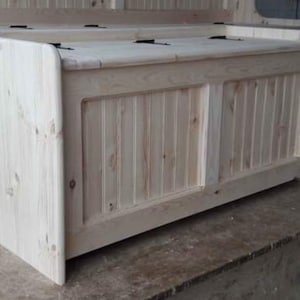 May include: A natural wood storage bench with a hinged lid and a panelled front. The bench is made of unfinished pine wood and has a simple, rustic design.