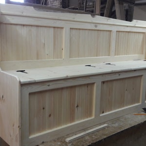 May include: A light-coloured wooden bench with a high backrest and vertical paneling details. The seat features three distinct sections with visible black metal hinges, indicating a storage compartment design. The wood grain is natural and unfinished.
