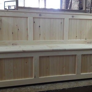 May include: A light-coloured wooden bench with a high backrest and a flat seat. The piece features a panelled design with vertical slats on both the backrest and the base, showcasing natural wood grain and knots throughout the light pine surface.