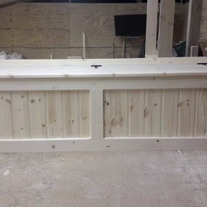 May include: A long, rectangular wooden storage bench with a natural light pine finish. The front panel features vertical beadboard detailing divided into two sections. The top lid is flat and secured with three visible black metal hinges.