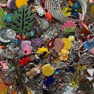 May include: A bowl filled with a variety of colourful charms and pendants. The charms feature diverse shapes, including leaves, flowers, animals, and food items. Many are silver-toned, with accents of yellow, pink, blue, and red. The bowl contains many small, decorative elements.