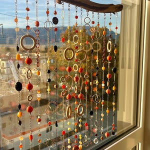 May include: A decorative window hanging with strands of beads in various shapes and colours, including orange, red, yellow, and black. The beads are suspended from a natural wooden branch, creating a unique and artistic display.
