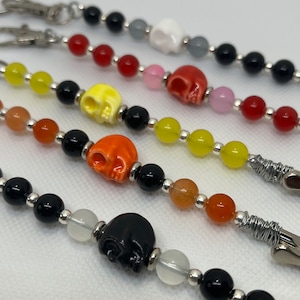 May include: Close-up of several beaded keychains. Each keychain features a different coloured skull bead, including white, red, yellow, orange, and black. The beads are strung with smaller beads in various colours and silver-toned metal accents.