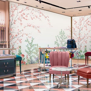 May include: A retail store interior with a floral wallpaper featuring pink blossoms and green leaves. The store has a black and white checkered floor, a green chest of drawers, and two pink and red chairs.