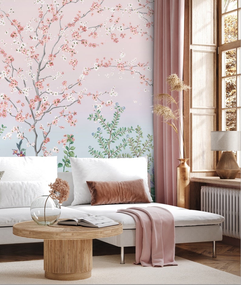 May include: A white couch with pink throw blankets and pillows sits in front of a wall with a pink and blue floral wallpaper. A wooden coffee table with a glass vase and dried flowers sits in front of the couch.