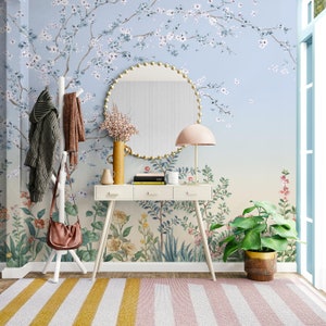 May include: A white entryway table with three drawers and a round mirror with a gold frame. The table is decorated with a vase of flowers, a book, and a candle. The wall behind the table is covered in a floral wallpaper with a blue background. A brown leather bag hangs on a white coat rack. A green plant sits in a brown pot on the floor.