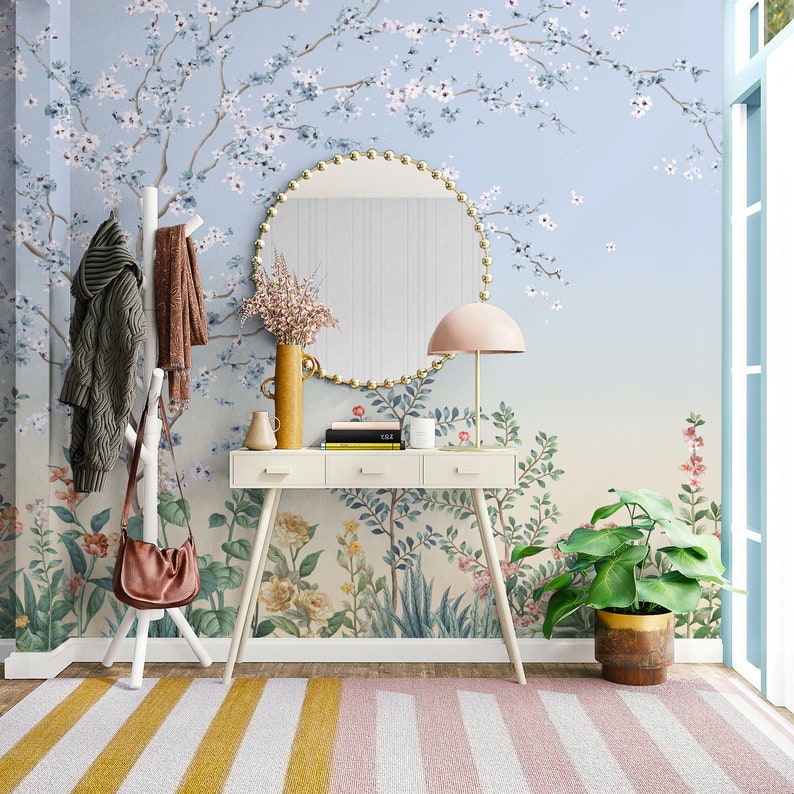 May include: A white entryway table with three drawers and a round mirror with a gold frame. The table is decorated with a vase of flowers, a book, and a candle. The wall behind the table is covered in a floral wallpaper with a blue background. A brown leather bag hangs on a white coat rack. A green plant sits in a brown pot on the floor.