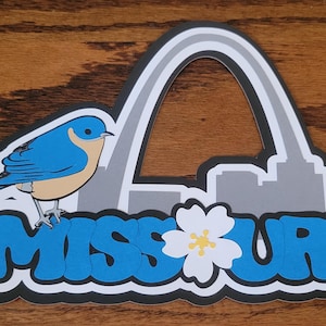 May include: A die-cut sticker featuring a bluebird perched above the word "MISSOURI" in blue bubble letters. The design includes the Gateway Arch and a white flower with a yellow center, all outlined in black.
