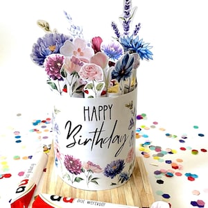 Puede incluir: Una tarjeta de cumpleaños cilíndrica blanca con la frase "HAPPY Birthday" en escritura negra, decorada con ilustraciones florales. La tarjeta está llena de flores de papel y se encuentra sobre una base de madera, rodeada de confeti colorido.