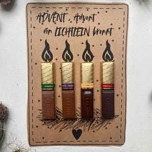 May include: A handmade Advent calendar card with four chocolate bars arranged to resemble candles. The card is brown with the German text "ADVENT, Advent ein LICHTLEIN brennt" and a heart. The chocolate bars have gold and colored wrappers.