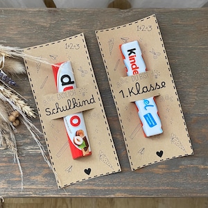 Puede incluir: Dos tarjetas de papel Kraft con barras de chocolate Kinder adjuntas. Las tarjetas tienen el texto "ABC ENDLICH Schulkind" y "ABC DU BIST 1. Klasse". Cada tarjeta tiene un borde punteado con dibujos de lápices, tijeras y zanahorias.