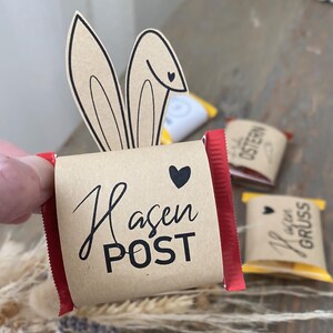 Könnte beinhalten: Eine rote und braune Schokoladentafel mit einer braunen Papierverpackung. Die Verpackung hat ein schwarzes Hasenohren-Design und den Text "Hasen POST" in Schwarz. Auf der Verpackung befindet sich ein kleines rotes Herz.