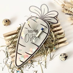 Könnte beinhalten: Eine karottenförmige Osterkarte mit den Worten "hoppelige Ostern" und einer aufgerollten Banknote im Inneren. Die Karte ist mit bunten Konfetti gefüllt und liegt auf einer Holzkonstruktion mit Heu und zwei gesprenkelten Eiern. Die Karotte hat einen schwarzen Umriss.