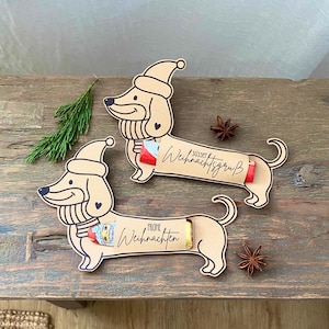 Puede incluir: Dos adornos navideños con forma de perro salchicha, hechos de cartón marrón claro. Cada adorno tiene un gorro de Papá Noel, una bufanda y contiene una pequeña chocolatina. El texto "Frohe Weihnachten" y "Süßer Weihnachtsgruß" está escrito.