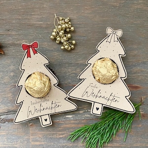 May include: Two Christmas tree-shaped gift holders made of light brown cardboard, each holding a gold-wrapped chocolate. One has a red bow and the text "zauberhafte Weihnachten", the other has a bow and the text "Frohe Weihnachten".