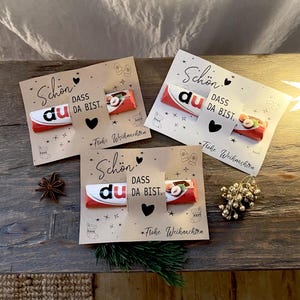以下が含まれることがあります： チョコレートバー付きのクリスマスカード3枚。各カードには「Schön dass du da bist」と「Frohe Weihnachten」の文字が印刷されています。カードはハート、星、お祝いのアイコンで飾られています。