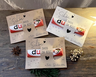 May include: Three Christmas cards with chocolate bars. Each card has the words "Schön dass du da bist" and "Frohe Weihnachten" printed on them. The cards are decorated with hearts, stars, and festive icons.
