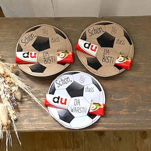 Puede incluir: Tres tarjetas circulares de papel con gráficos de balón de fútbol y texto. Dos tarjetas son de color canela con patrones negros y el texto "Schön, dass du da bist!" con un icono de signo de paz. Una tarjeta blanca tiene patrones negros y el texto "Schön, dass du warst!!". Cada tarjeta sostiene una pequeña barra de chocolate.