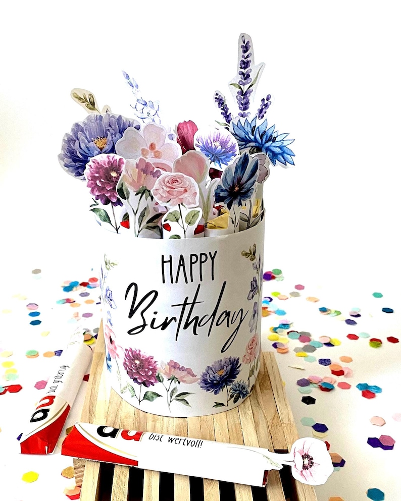 Pu&ograve; includere: Un biglietto di compleanno cilindrico bianco con la scritta "HAPPY Birthday" in corsivo nero, decorato con illustrazioni floreali. Il biglietto &egrave; pieno di fiori di carta e poggia su una base di legno, circondato da coriandoli colorati.
