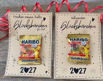 May include: Two rectangular tan cards with red ribbons, each featuring a small Haribo Goldbären gummy candy bag attached. The left card reads Frohes neues Jahr Glücksbärchen für dich 2027, and the right card reads Silvester Glücksbärchen 2027.