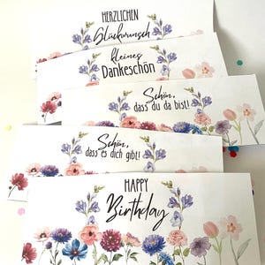 Pu&ograve; includere: Un gruppo di biglietti di auguri bianchi con disegni floreali e vari messaggi. Le carte presentano testi come "Happy Birthday" e frasi tedesche, circondati da fiori ad acquerello nei toni del rosa, blu e viola.