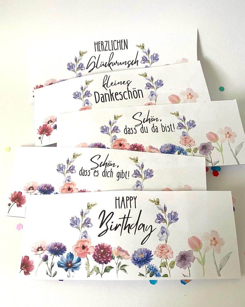 Pu&ograve; includere: Un gruppo di biglietti di auguri bianchi con disegni floreali e vari messaggi. Le carte presentano testi come "Happy Birthday" e frasi tedesche, circondati da fiori ad acquerello nei toni del rosa, blu e viola.