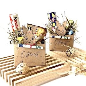 Può includere: Due piccole scatole di cartone marrone piene di dolcetti pasquali, tra cui barrette di cioccolato e uova di cioccolato. Ogni scatola ha un ritaglio di coniglietto e la parola "Osternest" stampata sul davanti. Un tema pasquale decorativo.