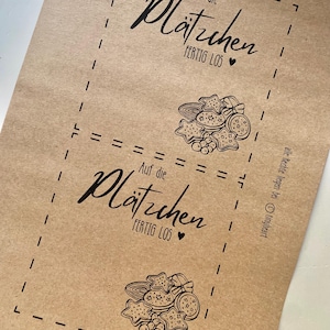 May include: Brown paper with two printed sections featuring the text "Auf die Pl&auml;tzchen FERTIG LOS" and a heart symbol. Each section includes a drawing of various cookies.