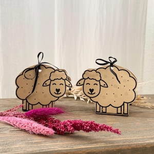 Puede incluir: Dos cajas de regalo de papel marrón con forma de oveja con lazos de cinta negra. Las ovejas tienen ojos, narices y bocas negras, y manchas blancas en sus cuerpos. Las cajas están sobre una superficie de madera con flores secas rosas.