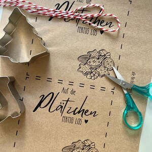 May include: A kraft paper sheet with printed cookie designs, text, and dotted lines. A red and white twine and a pair of scissors with teal handles are on the paper. Metal cookie cutters in the shape of a Christmas tree and a star are also present.