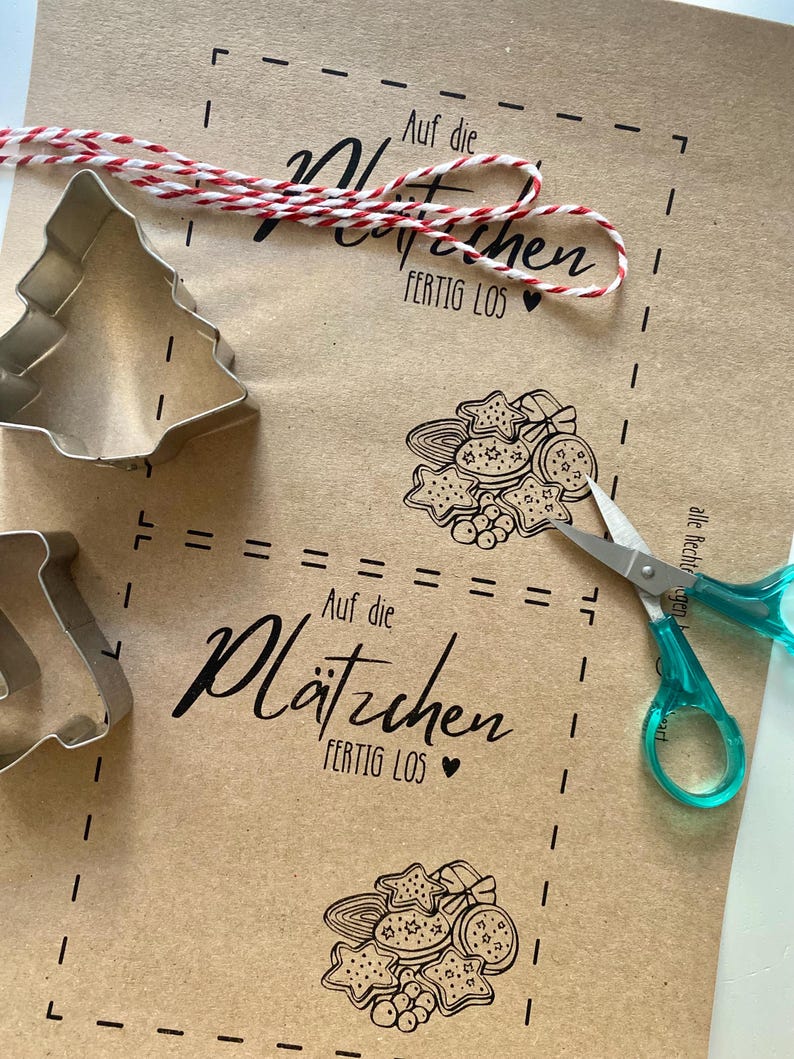 May include: A kraft paper sheet with printed cookie designs, text, and dotted lines. A red and white twine and a pair of scissors with teal handles are on the paper. Metal cookie cutters in the shape of a Christmas tree and a star are also present.
