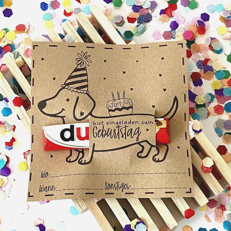 Puede incluir: Una tarjeta marr&oacute;n con una ilustraci&oacute;n de un perro salchicha con un gorro de fiesta y sosteniendo un pastel. La tarjeta tiene las palabras "du" y "Geburtstag", con espacio para escribir "Wo" y "Wann".