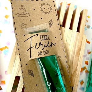 May include: A kraft paper card with summer-themed illustrations and the text "COOLE Ferien F&Uuml;R DICH" is attached to a wooden structure. The card holds a green ice pop. Additional ice pops are visible in the background.