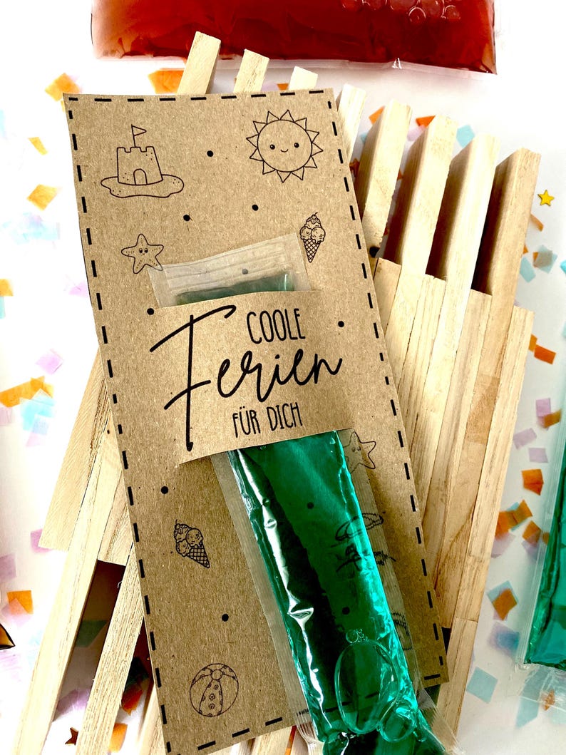 May include: A kraft paper card with summer-themed illustrations and the text "COOLE Ferien F&Uuml;R DICH" is attached to a wooden structure. The card holds a green ice pop. Additional ice pops are visible in the background.
