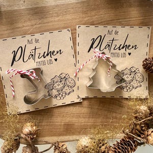 May include: Two kraft paper cards with cookie cutter shapes and the text "Auf die Pl&auml;tzchen fertig los". One card has a stocking-shaped cutter, the other a Christmas tree. Both are tied with red and white twine.