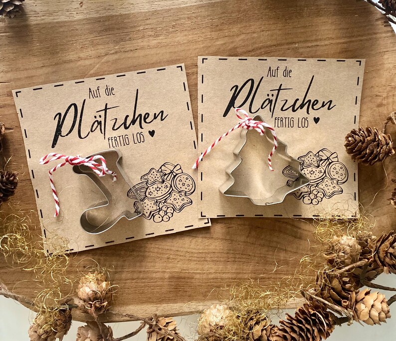 May include: Two kraft paper cards with cookie cutter shapes and the text "Auf die Pl&auml;tzchen fertig los". One card has a stocking-shaped cutter, the other a Christmas tree. Both are tied with red and white twine.