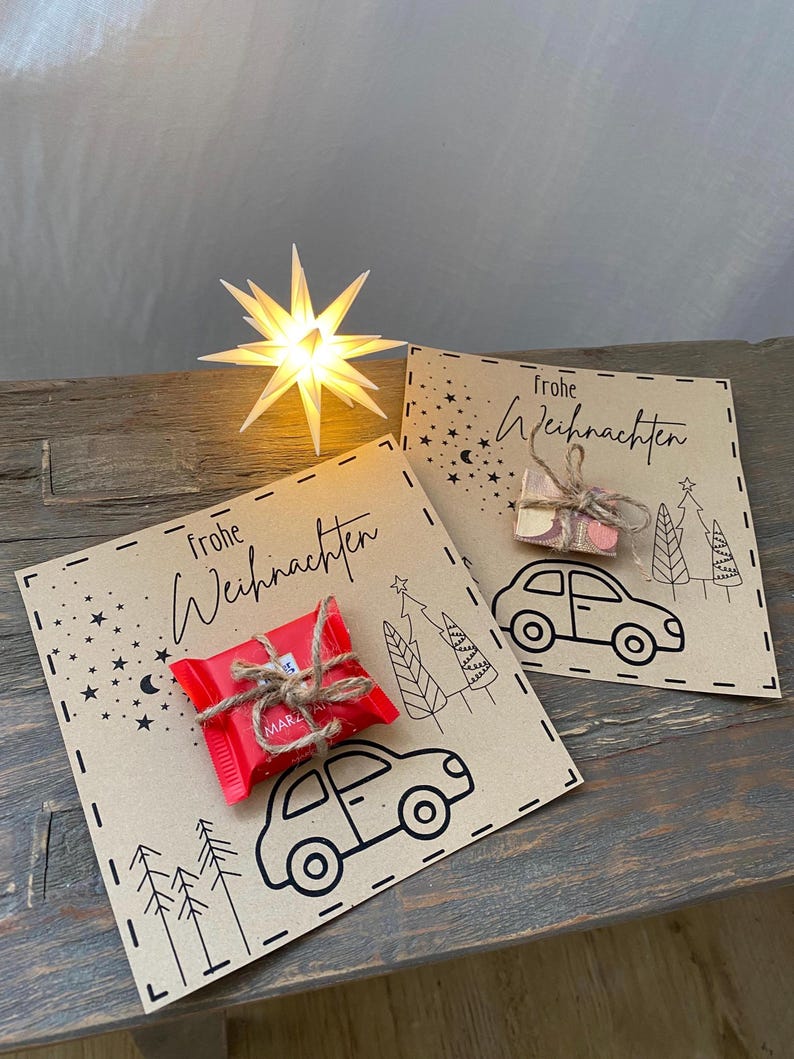 May include: Two kraft paper cards with the words "Frohe Weihnachten" and a car illustration. One card has a red wrapped candy tied with twine. The other card has a small bag of pink and white candies. A lit star-shaped decoration is in the background.