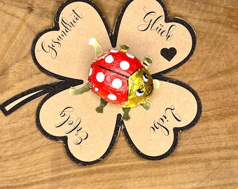 May include: A red and white ladybug-shaped chocolate candy sits atop a four-leaf clover-shaped card. The card is tan with black edging and features the words "Gesundheit", "Glück", "Erfolg", and "Liebe". The background is a wooden surface.