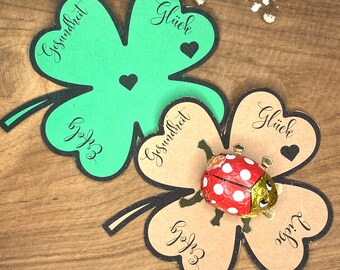 May include: Two four-leaf clovers, one green and one brown, with the words "Glück" and "Gesundheit" written on them. A small red ladybug chocolate is on top of the brown clover.