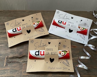 May include: Three greeting cards with a Kinder chocolate bar attached. The cards are decorated with the words "Schön dass du da bist" and "Frohes neues Jahr", along with hearts and starbursts. The chocolate bars have red and white packaging.