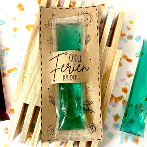 May include: A green gel pack in a clear plastic pouch, presented on a brown card with the text "COOLE Ferien F&Uuml;R DICH". The card is decorated with beach-themed illustrations. The gel pack is displayed on a wooden structure.