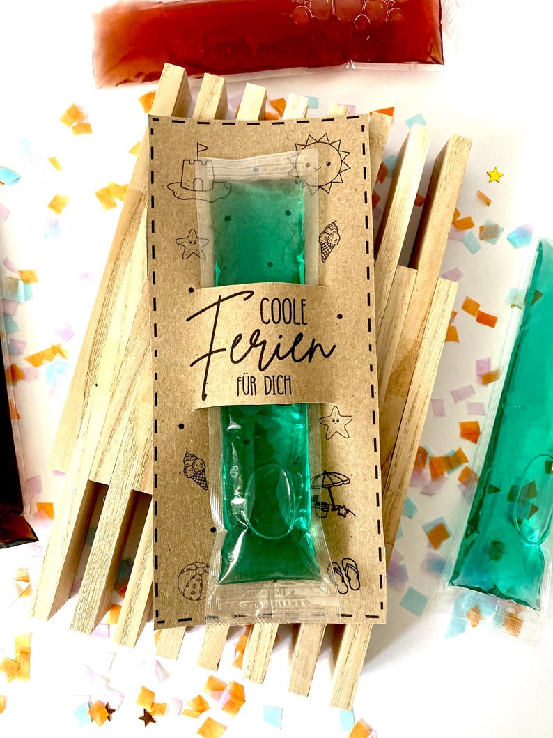 May include: A green gel pack in a clear plastic pouch, presented on a brown card with the text "COOLE Ferien F&Uuml;R DICH". The card is decorated with beach-themed illustrations. The gel pack is displayed on a wooden structure.