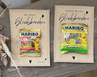 May include: Two cards with Haribo gummy bear candies. The cards have the text "ein paar Glücksbärcher" and "nimm dir ein Glücksbärcher" in black script. The Haribo packages are gold and green, with the Haribo logo and the words "Goldbären".