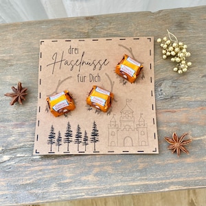 Op de afbeelding: Een vierkante bruine kaart met de tekst "drei Haselnüsse für Dich", een lijntekening van een kasteel en dennenbomen. Drie oranje verpakte Ferrero Küsschen chocolaatjes zijn op de kaart bevestigd, samen met twee steranijs en een gouden bessentak.