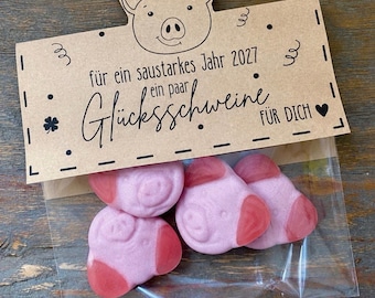 May include: A clear plastic bag containing four pink pig-shaped gummy candies. The bag is sealed with a brown paper topper featuring a line drawing of a pig face, a four-leaf clover, a heart, and the text 'für ein saustarkes Jahr 2027 ein paar Glücksschweine FÜR DICH'.