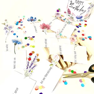 Pu&ograve; includere: Una tazza bianca con un motivo floreale e la scritta "HAPPY Birthday" &egrave; posizionata vicino a coriandoli sparsi e piccole carte con illustrazioni floreali e testo in tedesco. Le forbici sono visibili, suggerendo un tema artigianale o di regalo.