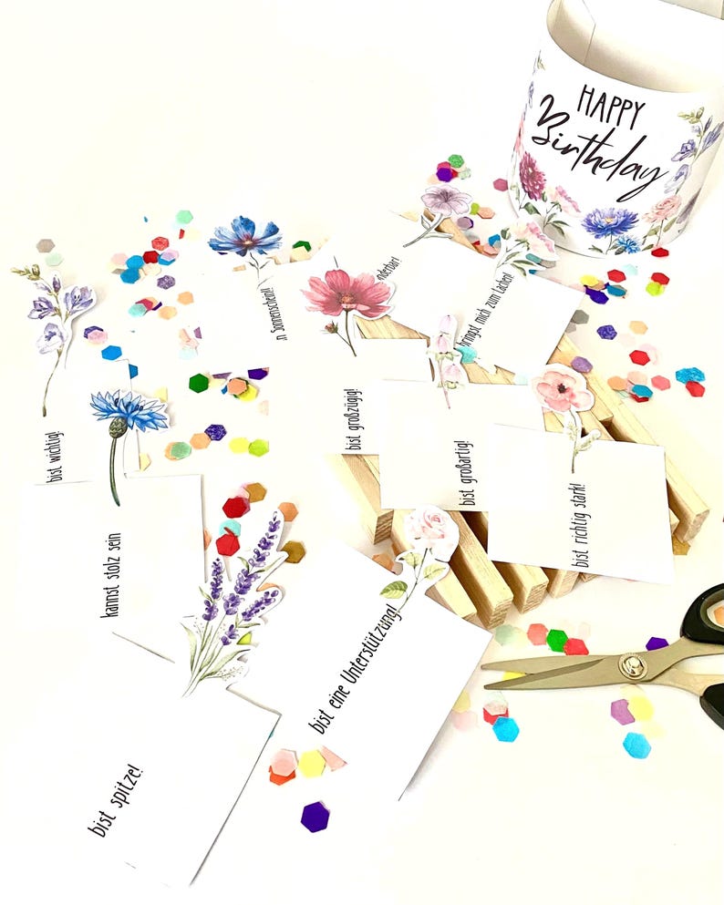 Pu&ograve; includere: Una tazza bianca con un motivo floreale e la scritta "HAPPY Birthday" &egrave; posizionata vicino a coriandoli sparsi e piccole carte con illustrazioni floreali e testo in tedesco. Le forbici sono visibili, suggerendo un tema artigianale o di regalo.