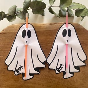 Könnte beinhalten: Zwei weiße, geisterförmige Halloween-Dekorationen mit schwarzen Umrissen, schwarzen ovalen Augen und dem Wort "Boo!" in geschwungener Schrift. Jeder Geist hat einen leuchtend orangefarbenen und rosa Strohhalm eingesetzt. Grüne Blätter sind im Hintergrund.