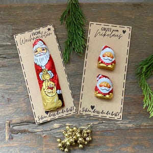 Könnte beinhalten: Zwei Schokoladen-Weihnachtsmänner in roter und goldener Folie, präsentiert auf braunen Karten mit deutschem Text und Stern- und Herzdesign. Das Bild enthält einen Zweig Grün und goldene Glocken, für Weihnachten.